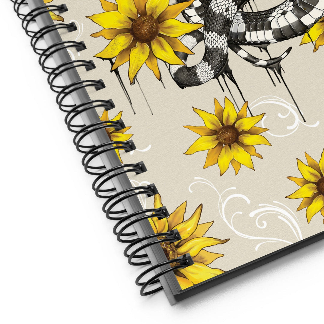A close-up of a spiral-bound notebook showing the cover design, which features yellow sunflowers and a black-and-white snake on a light beige background.