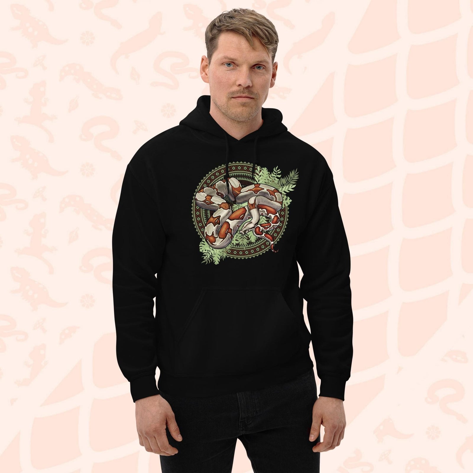 A man wearing a black hooded sweatshirt with a graphic print of a red tailed boa constrictor snake with bold red's and brown markings. The snake is curled up in a circle made of patterned geometric shapes and with leafy green foliage beneath the snakes body. The hoodie has a front pocket.