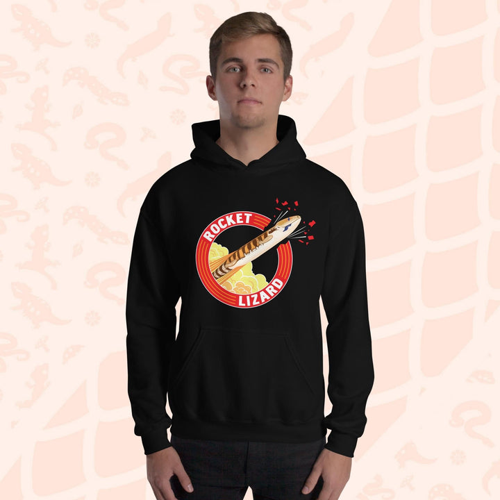 A man wearing a black hooded sweatshirt with a red circle graphic with text saying "Rocket Lizard." In the center of the graphic is a cartoon blue tongue skink shooting upward and breaking through the red outer circle with the text. It's tongue is hanging out of it's mouth as it rockets out with a plume of yellow and orange clouds billowing behind it. The hoodie has a front pocket.