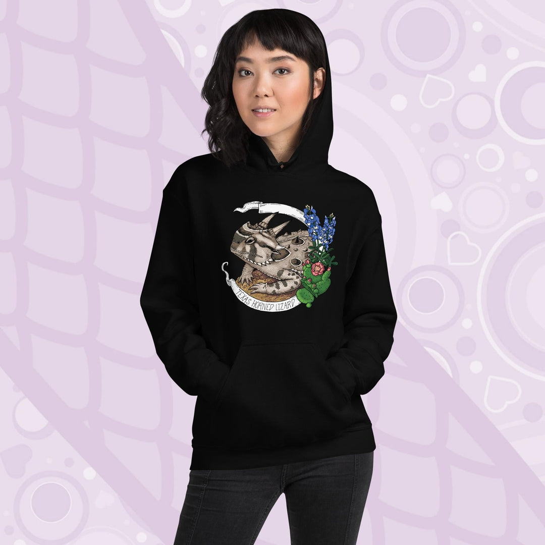 A woman wearing a black hoodie with a graphic of a Texas horned lizard surrounded by bluebonnets and a cactus. The text "Texas Horned Lizard" is printed in a white banner. The hoodie has a pocket in the front.