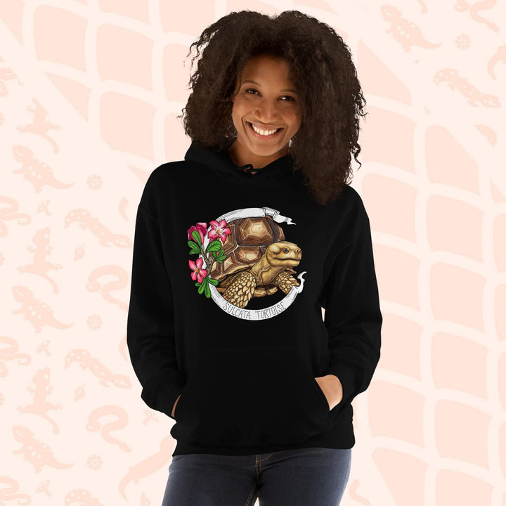A woman wearing a black hooded sweatshirt with a graphic of a Sulcata tortoise with large pink flowers on the left and a white banner framing above and below the artwork. Text "Sulcata Tortoise" is printed on the lower part of the banner. The hoodie has a front pouch.