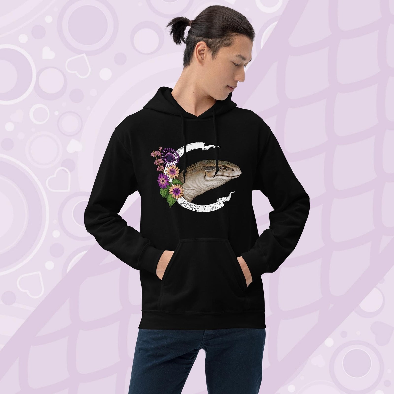 A  man wearing black hooded sweatshirt with a graphic of an African Savannah monitor lizard with red flowers and the text "Savannah Monitor" on the bottom white banner that frames the reptile. The hoodie is a cotton fabric and has a front pouch.