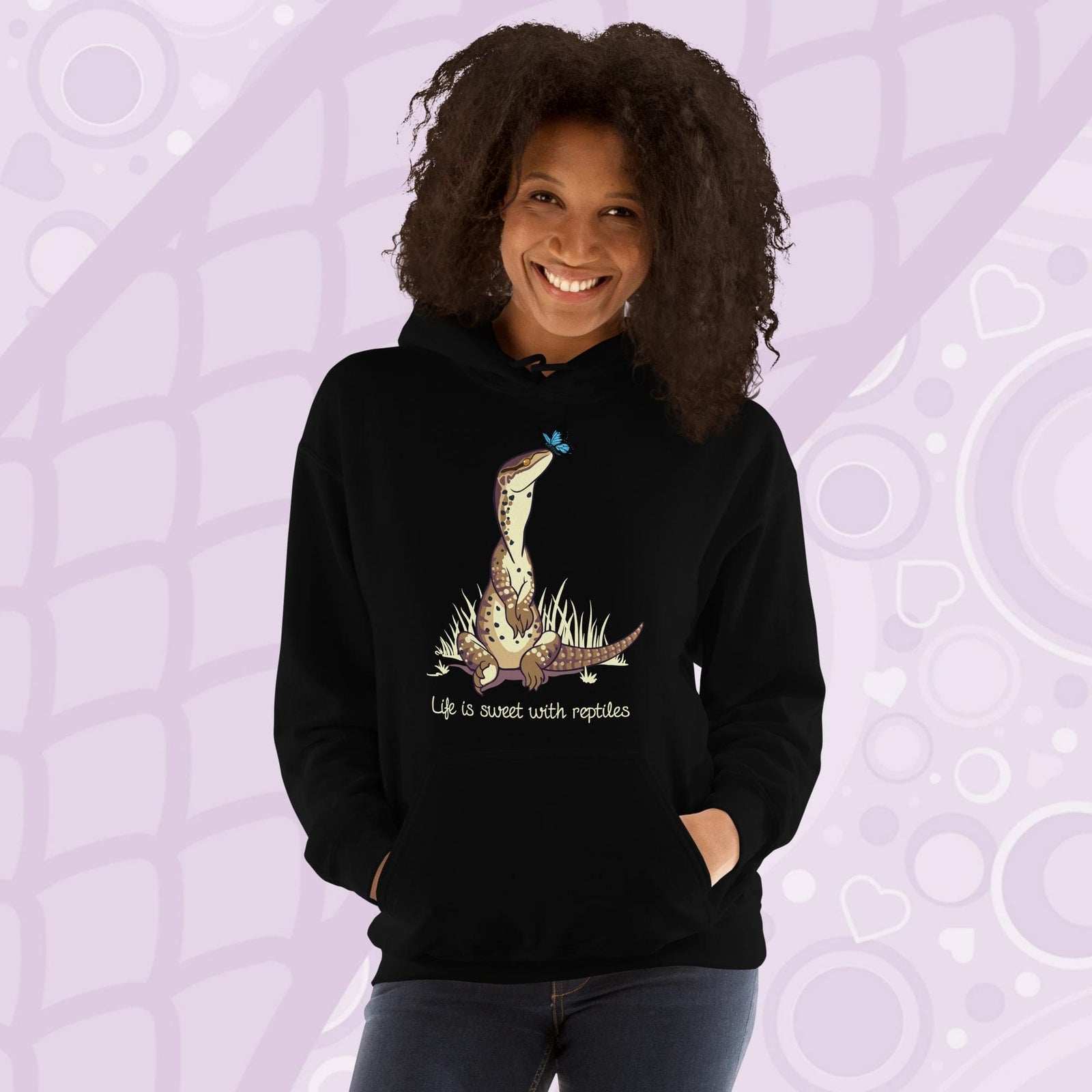 A smiling woman wearing a black hoodie with a front pouch and a cartoon graphic on the front of an argus monitor lizard sitting in grass with a little butterfly on it's nose. The text underneath says, "Life is sweet with reptiles."