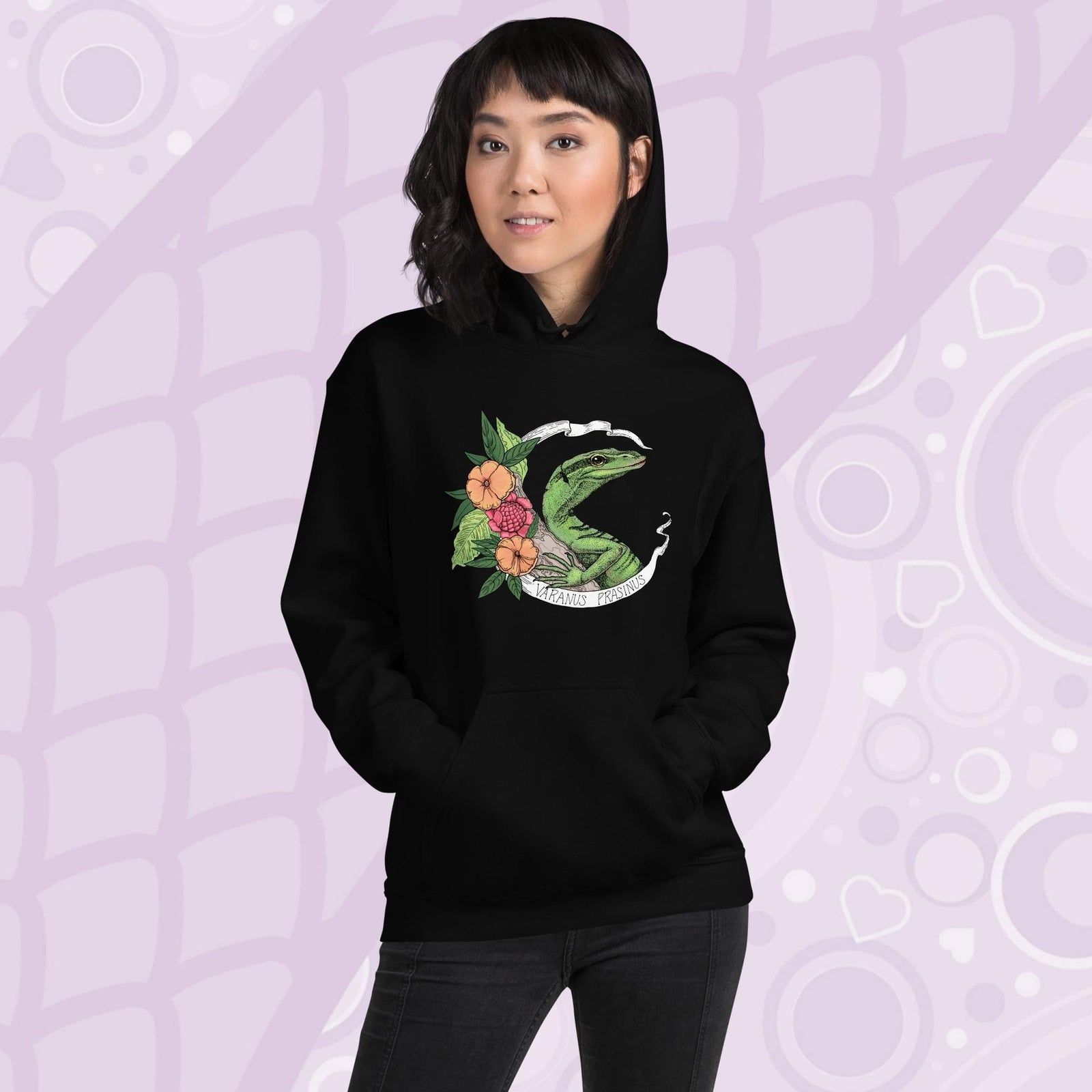 A woman wearing a black hoodie with a graphic of a slender green tree monitor lizard with orange and warm orange red flowers and the text "Varanus Prasinus" on a white banner surrounding the reptile. The hoodie has a front pocket.