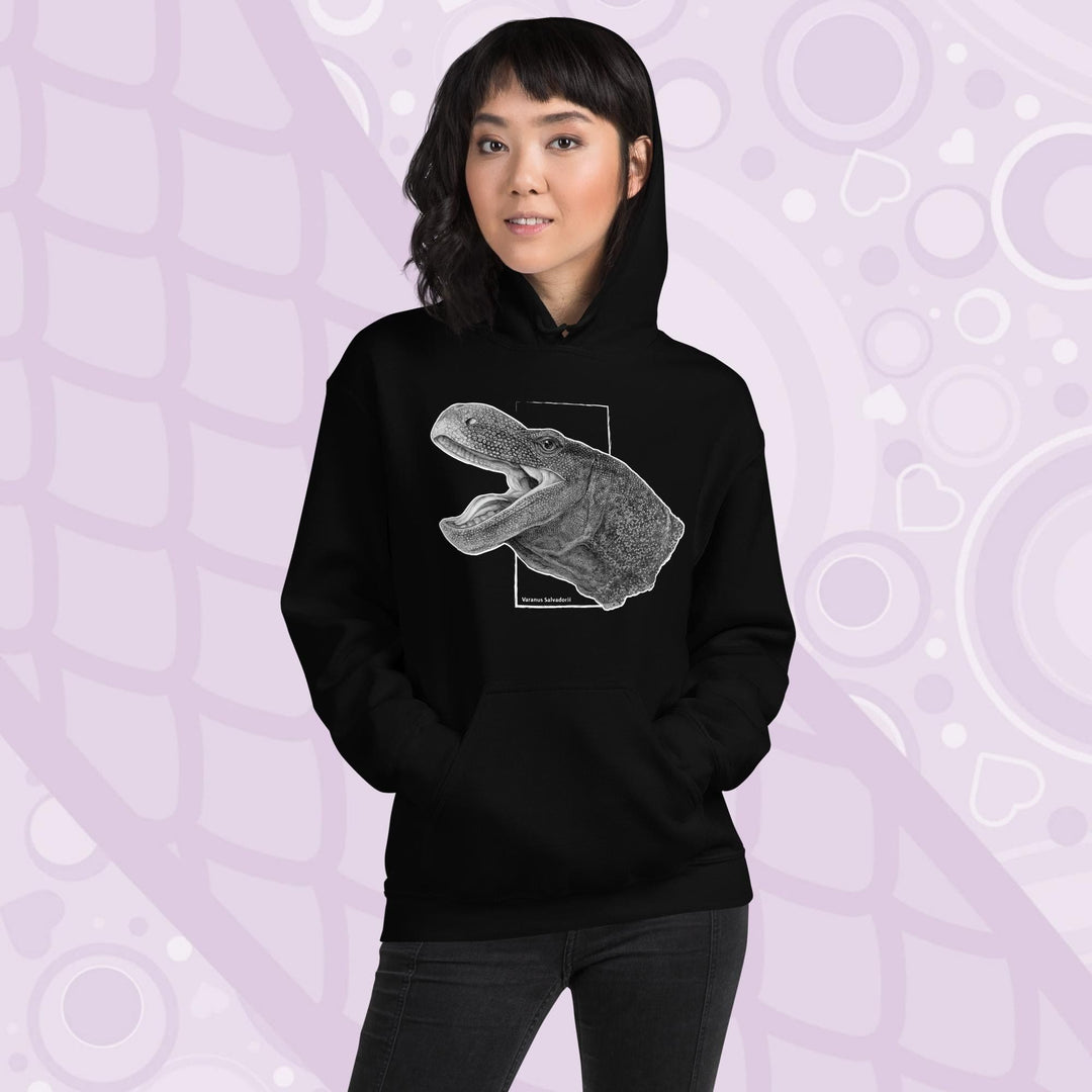 A woman wearing a black hooded sweatshirt with a graphite illustration of a head portrait of a crocodile monitor reptile with its mouth open. The illustration is framed by a white rectangle and the text "Varanus Salvadorii" is below the image. The hoodie has a front pocket.