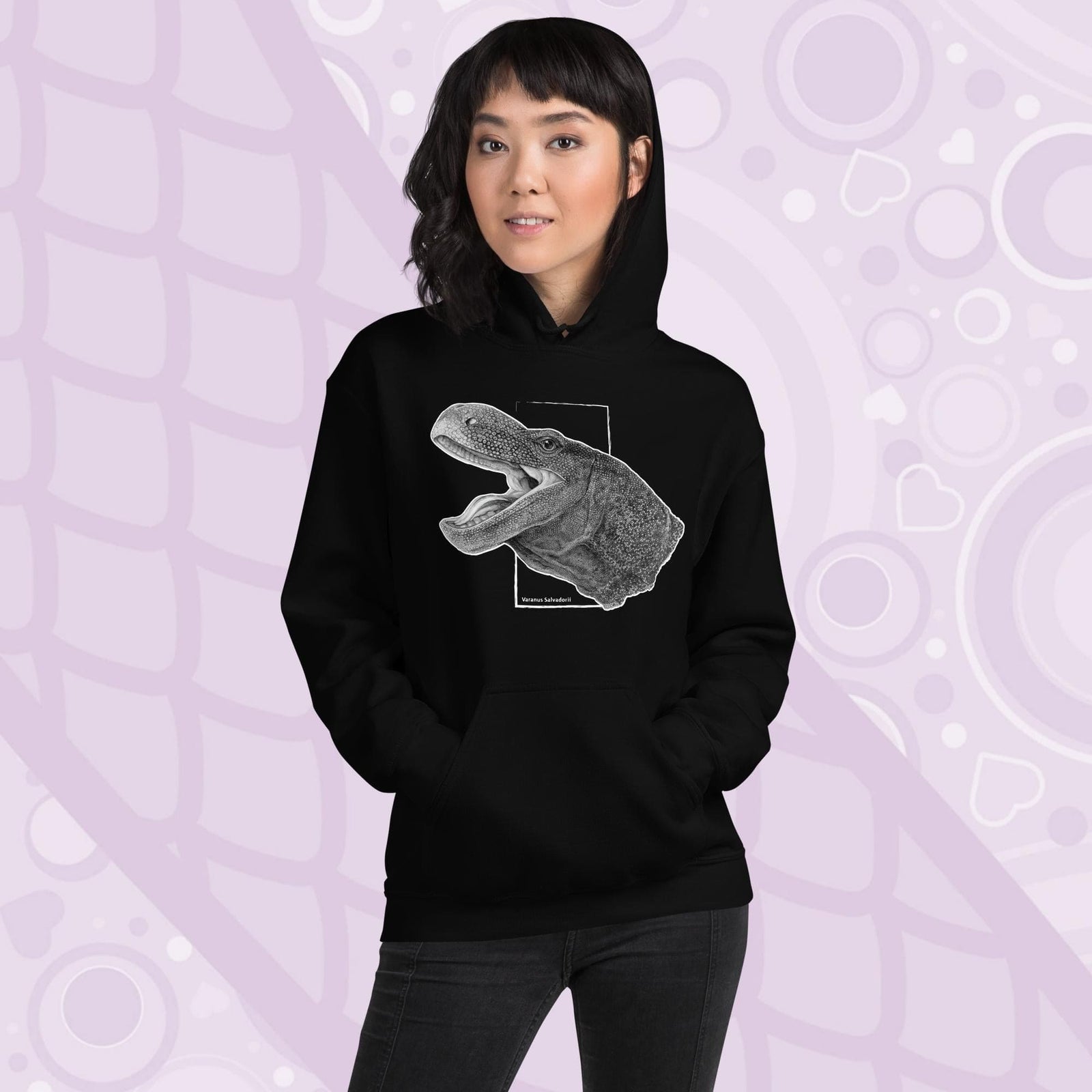 A woman wearing a black hooded sweatshirt with a graphite illustration of a head portrait of a crocodile monitor reptile with its mouth open. The illustration is framed by a white rectangle and the text "Varanus Salvadorii" is below the image. The hoodie has a front pocket.