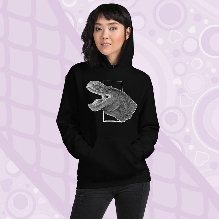 A woman wearing a black hooded sweatshirt with a graphite illustration of a head portrait of a crocodile monitor reptile with its mouth open. The illustration is framed by a white rectangle and the text "Varanus Salvadorii" is below the image. The hoodie has a front pocket.