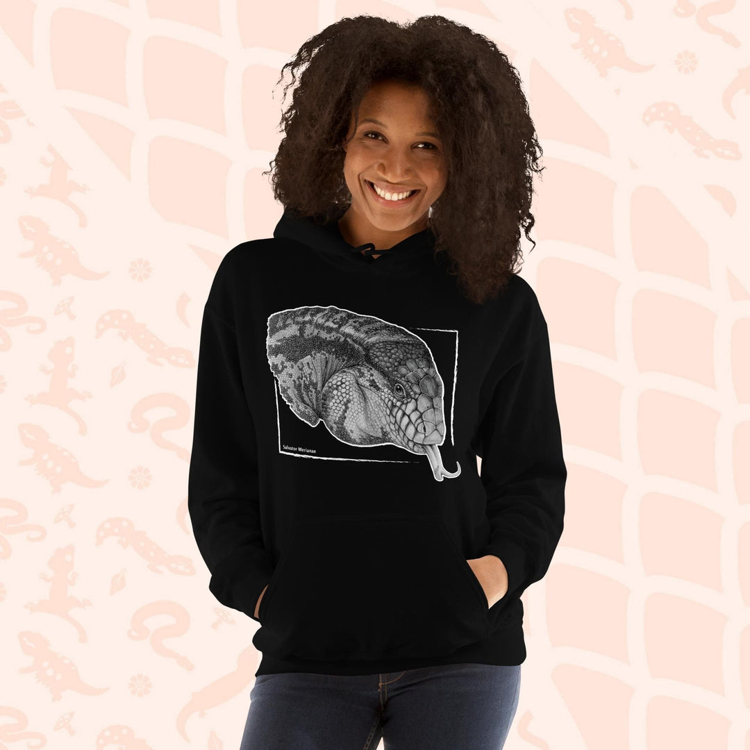 A woman wearing a black hooded sweatshirt featuring a detailed black and white illustration of an Argentine Tegus head with its tongue extended. The artwork is framed within a white rectangle with the reptiles species name of "salvator merianae" on the bottom. The hoodie has a front pocket on it.
