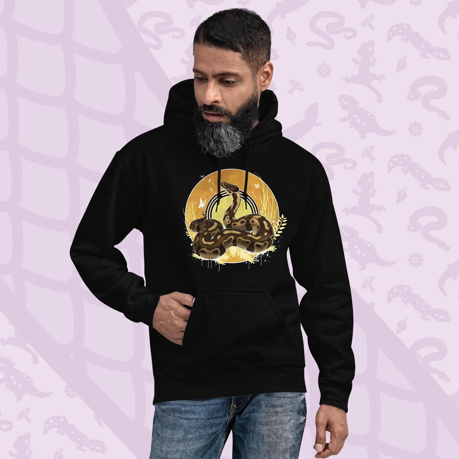Black hoodie with a graphic of a normal ball python snake with it's amber and brown colored scales coiled in a circle with it's head lifted up and looking upward. Behind it is a series of circles with golden hues and tall amber grasses with white butterflies and seeds floating on the wind.