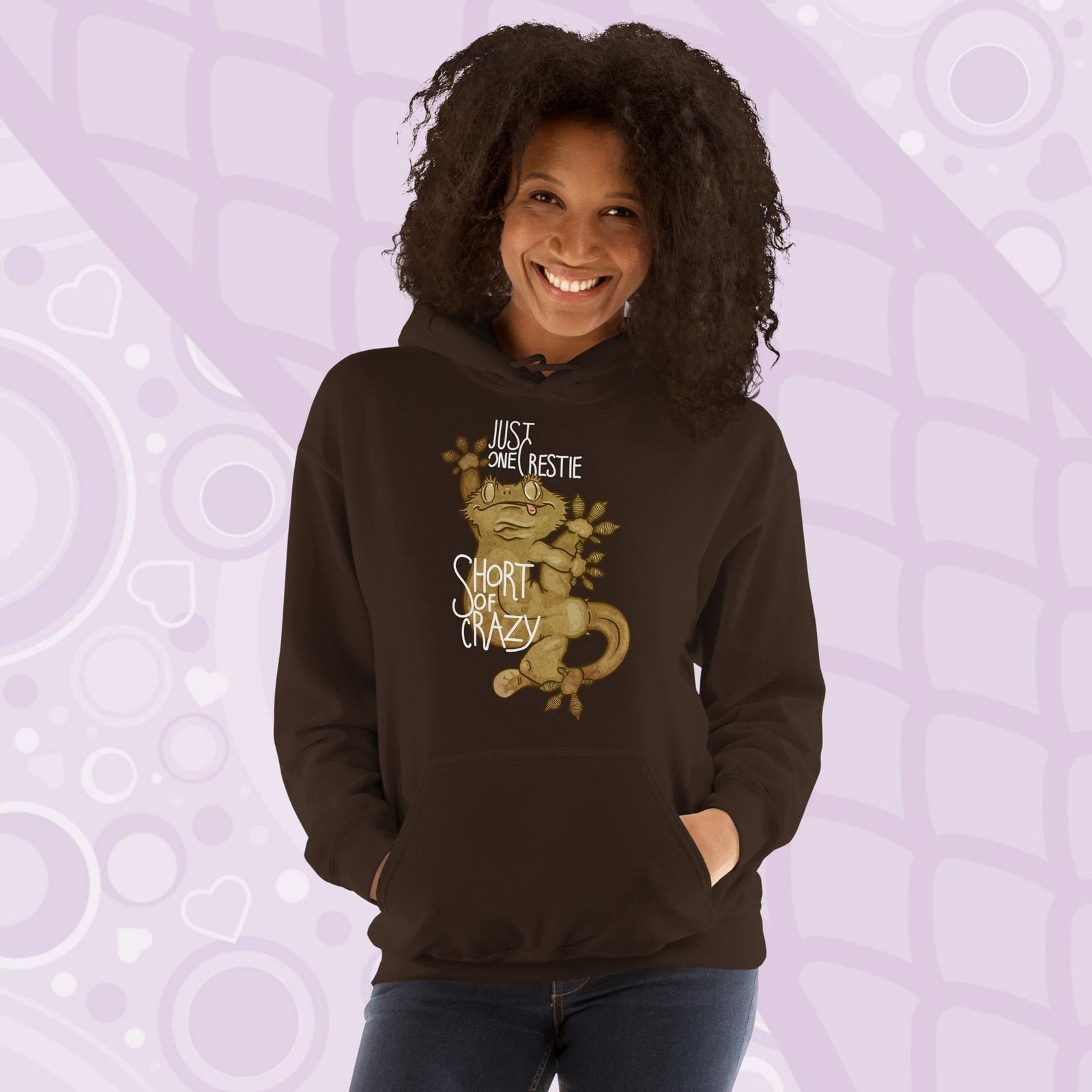 Woman wearing a brown colored pullover hoodie with a goofy, cartoonish graphic of a crested gecko with it's legs contorted in many directions. The text, "Just one crestie short of crazy." is printed on the front.