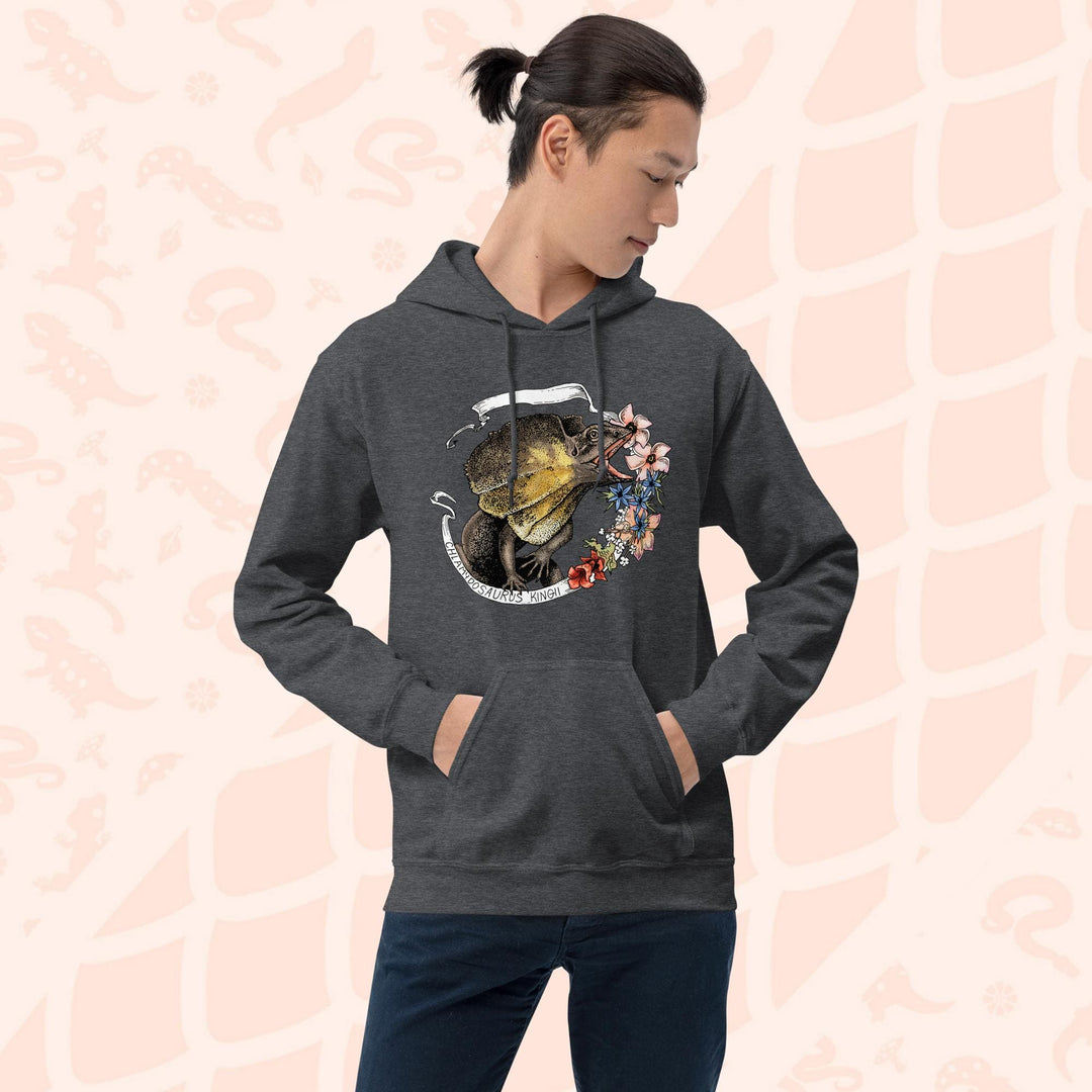Man wearing a grey hooded sweatshirt with a graphic of a frilled lizard, it's mouth open and displaying it's pink and orange frills with a white banner framing it's shape. Pink flowers flank the right side of the design and the text "Chlamydosaurus Kingii" is along the bottom, which is the species name for frilled lizard. The hoodie has a front pocket.
