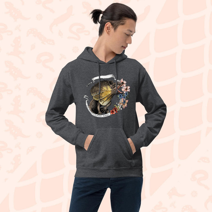 Man wearing a grey hooded sweatshirt with a graphic of a frilled lizard, it's mouth open and displaying it's pink and orange frills with a white banner framing it's shape. Pink flowers flank the right side of the design and the text "Chlamydosaurus Kingii" is along the bottom, which is the species name for frilled lizard. The hoodie has a front pocket.