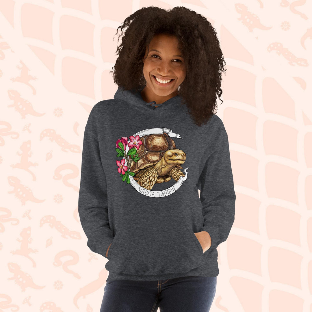 A woman wearing a grey hooded sweatshirt with a graphic of a Sulcata tortoise with large pink flowers on the left and a white banner framing above and below the artwork. Text "Sulcata Tortoise" is printed on the lower part of the banner. The hoodie has a front pouch.