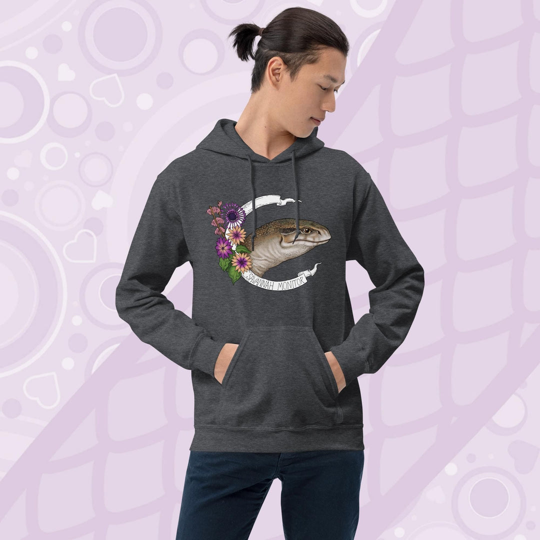 A  man wearing dark grey hooded sweatshirt with a graphic of an African Savannah monitor lizard with red flowers and the text "Savannah Monitor" on the bottom white banner that frames the reptile. The hoodie is a cotton fabric and has a front pouch.