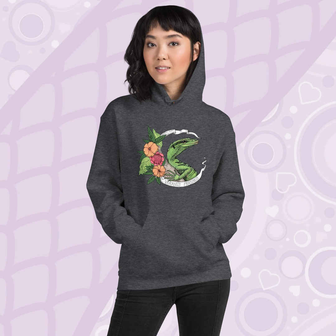 A woman wearing a dark heather grey hoodie with a graphic of a slender green tree monitor lizard with orange and warm orange red flowers and the text "Varanus Prasinus" on a white banner surrounding the reptile. The hoodie has a front pocket.