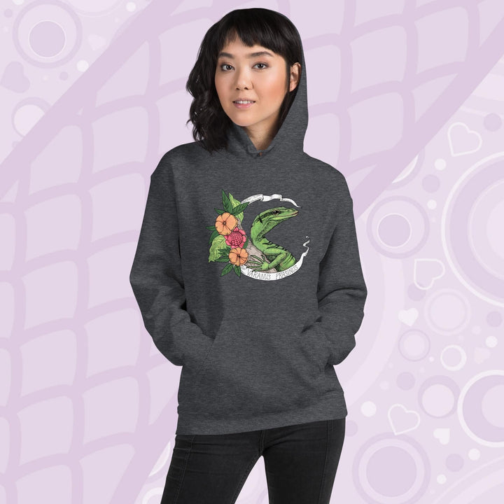 A woman wearing a dark heather grey hoodie with a graphic of a slender green tree monitor lizard with orange and warm orange red flowers and the text "Varanus Prasinus" on a white banner surrounding the reptile. The hoodie has a front pocket.