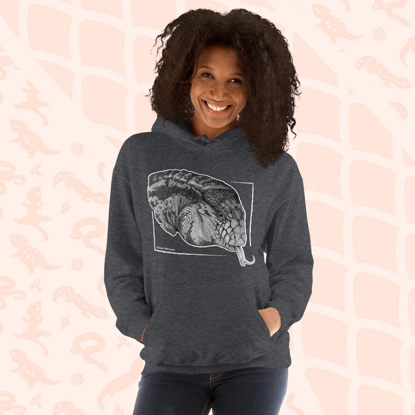 A woman wearing a grey hooded sweatshirt featuring a detailed black and white illustration of an Argentine Tegus head with its tongue extended. The artwork is framed within a white rectangle with the reptiles species name of "salvator merianae" on the bottom. The hoodie has a front pocket on it.