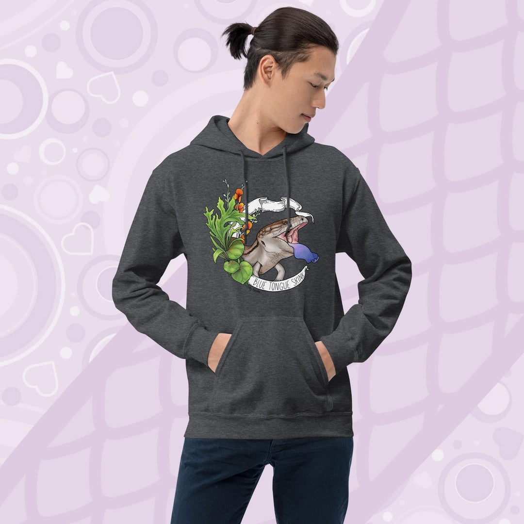 Man wearing a dark grey hooded pullover sweatshirt with a graphic of a blue-tongued skink, it's mouth open and it's blue tongue flashing in a classic defensive posture. The lizard is surrounded by green leaves and flowers. The text "Blue Tongue Skink" is written on a white banner.