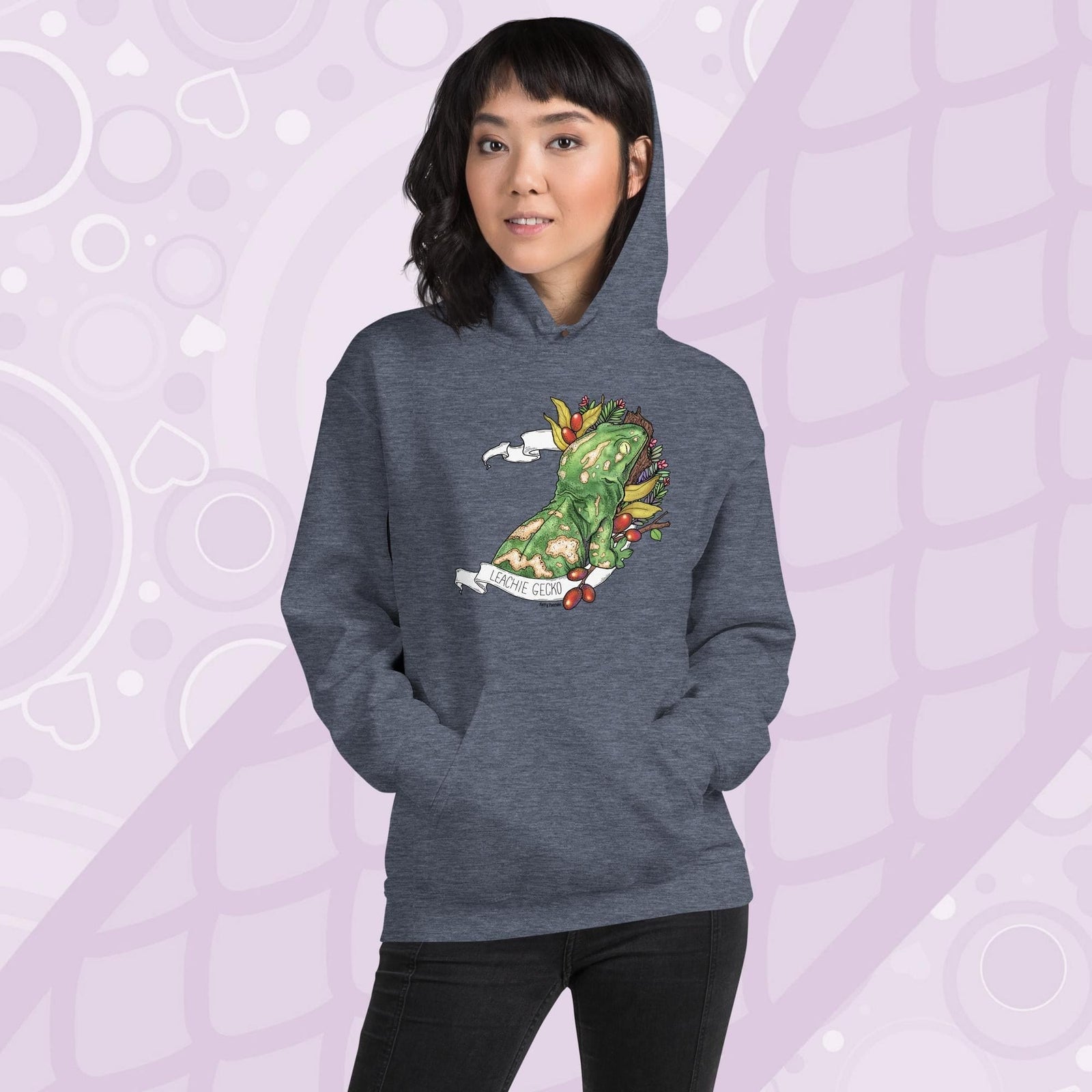 A woman wearing a grey pullover hoodie with a graphic of a leachie gecko leaning on a log framed by large green ferns and a white banner with the text "Leachie Gecko" on the bottom of the design. The hoodie is cotton material with a front pouch sewn in.