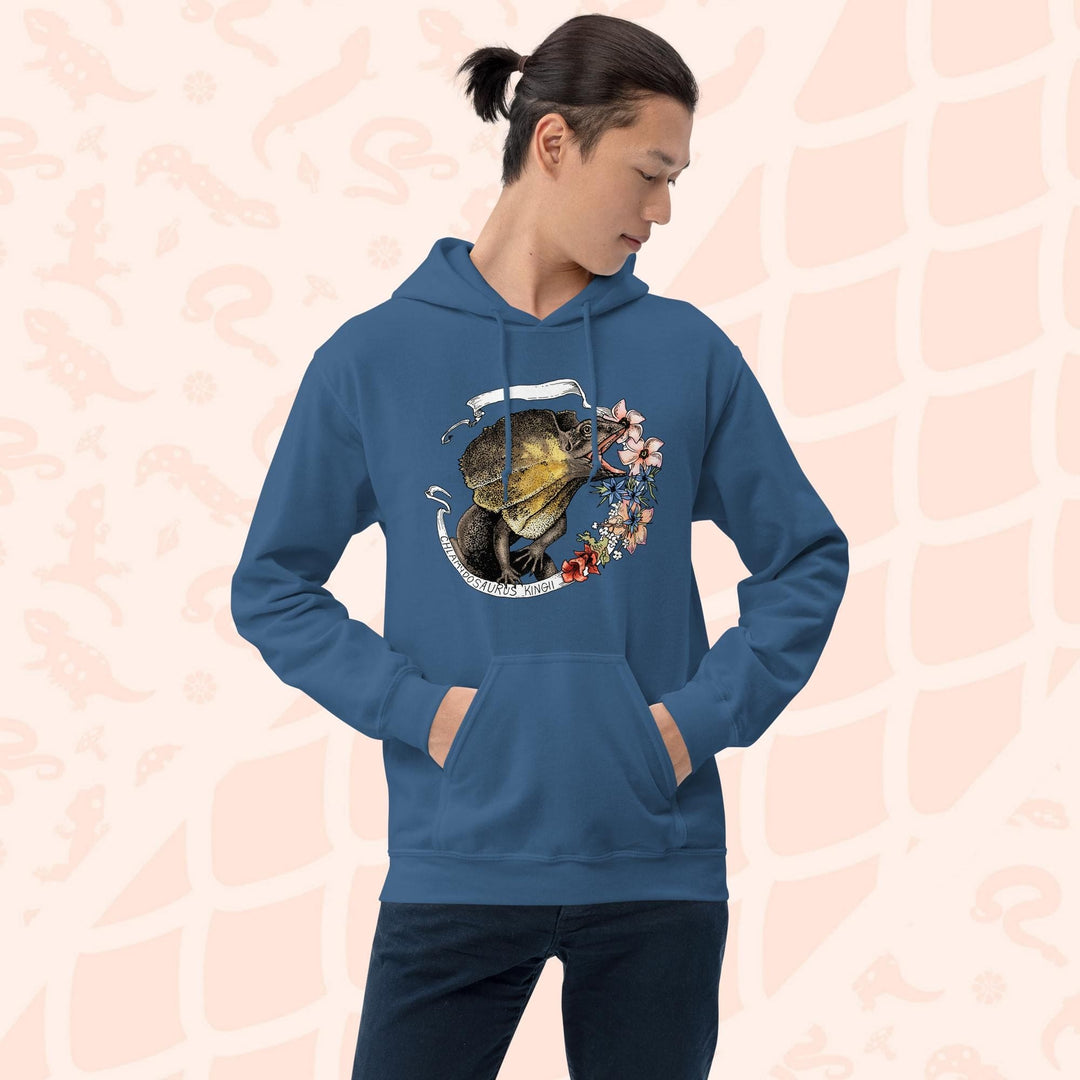 Man wearing a neutral blue hooded sweatshirt with a graphic of a frilled lizard, it's mouth open and displaying it's pink and orange frills with a white banner framing it's shape. Pink flowers flank the right side of the design and the text "Chlamydosaurus Kingii" is along the bottom, which is the species name for frilled lizard. The hoodie has a front pocket.