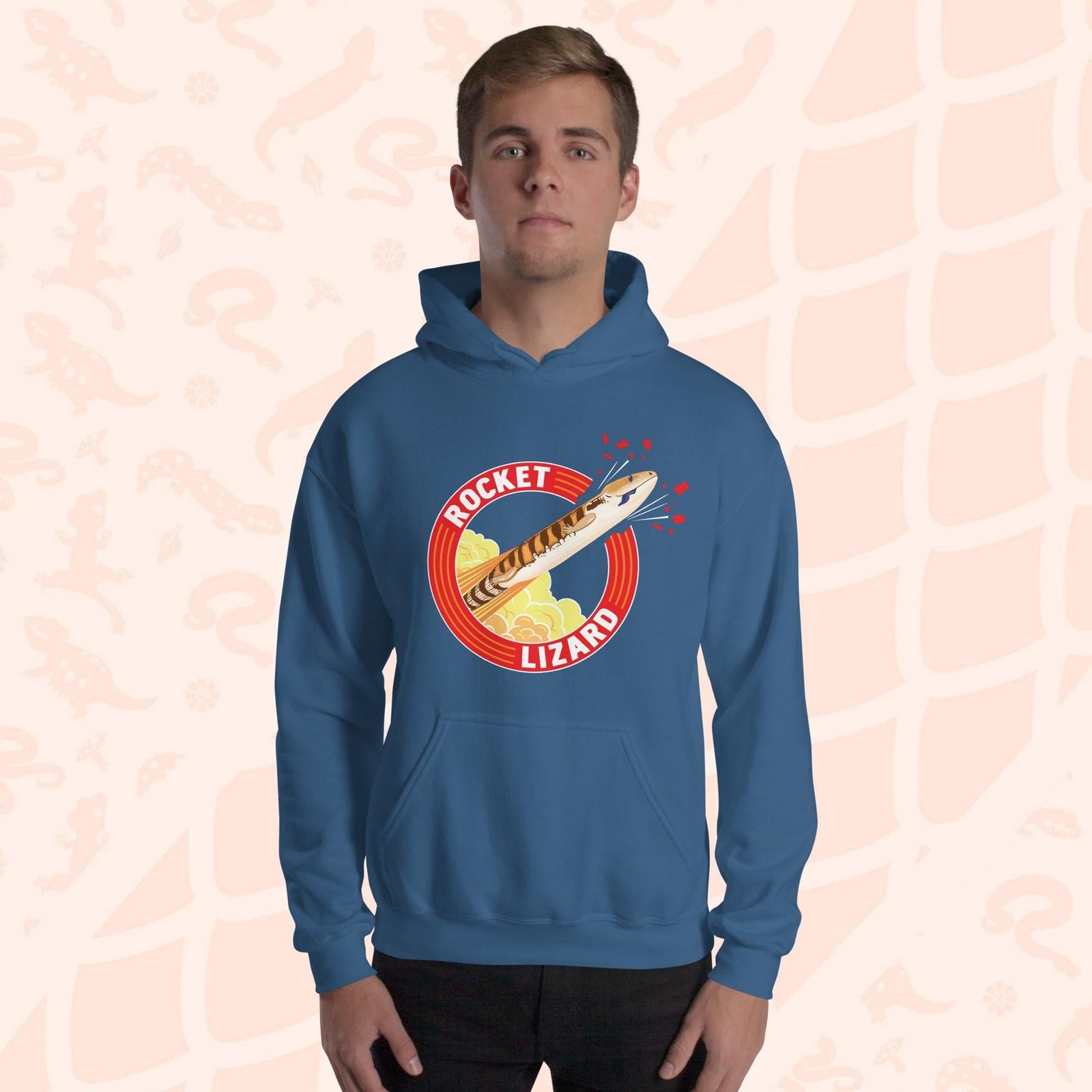 A man wearing a neutral blue hooded sweatshirt with a red circle graphic with text saying "Rocket Lizard." In the center of the graphic is a cartoon blue tongue skink shooting upward and breaking through the red outer circle with the text. It's tongue is hanging out of it's mouth as it rockets out with a plume of yellow and orange clouds billowing behind it. The hoodie has a front pocket.
