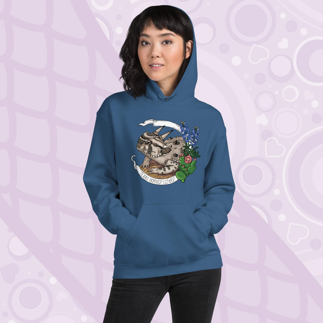 A woman wearing a neutral blue hoodie with a graphic of a Texas horned lizard surrounded by bluebonnets and a cactus. The text "Texas Horned Lizard" is printed in a white banner. The hoodie has a pocket in the front.