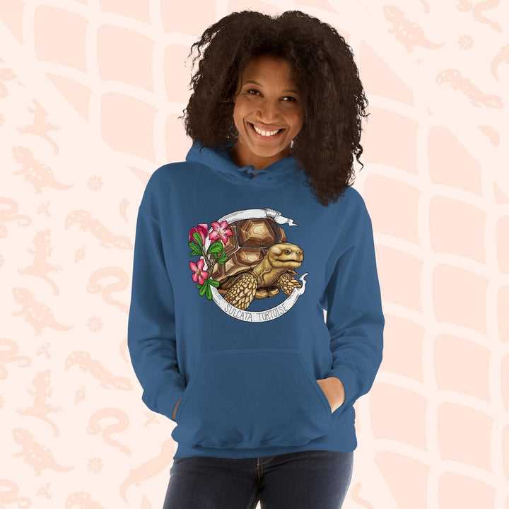 A woman wearing a neutral blue hooded sweatshirt with a graphic of a Sulcata tortoise with large pink flowers on the left and a white banner framing above and below the artwork. Text "Sulcata Tortoise" is printed on the lower part of the banner. The hoodie has a front pouch.