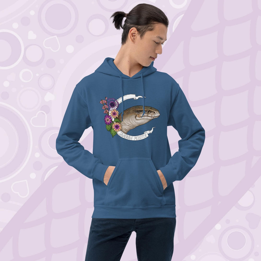 A  man wearing neutral blue hooded sweatshirt with a graphic of an African Savannah monitor lizard with red flowers and the text "Savannah Monitor" on the bottom white banner that frames the reptile. The hoodie is a cotton fabric and has a front pouch.