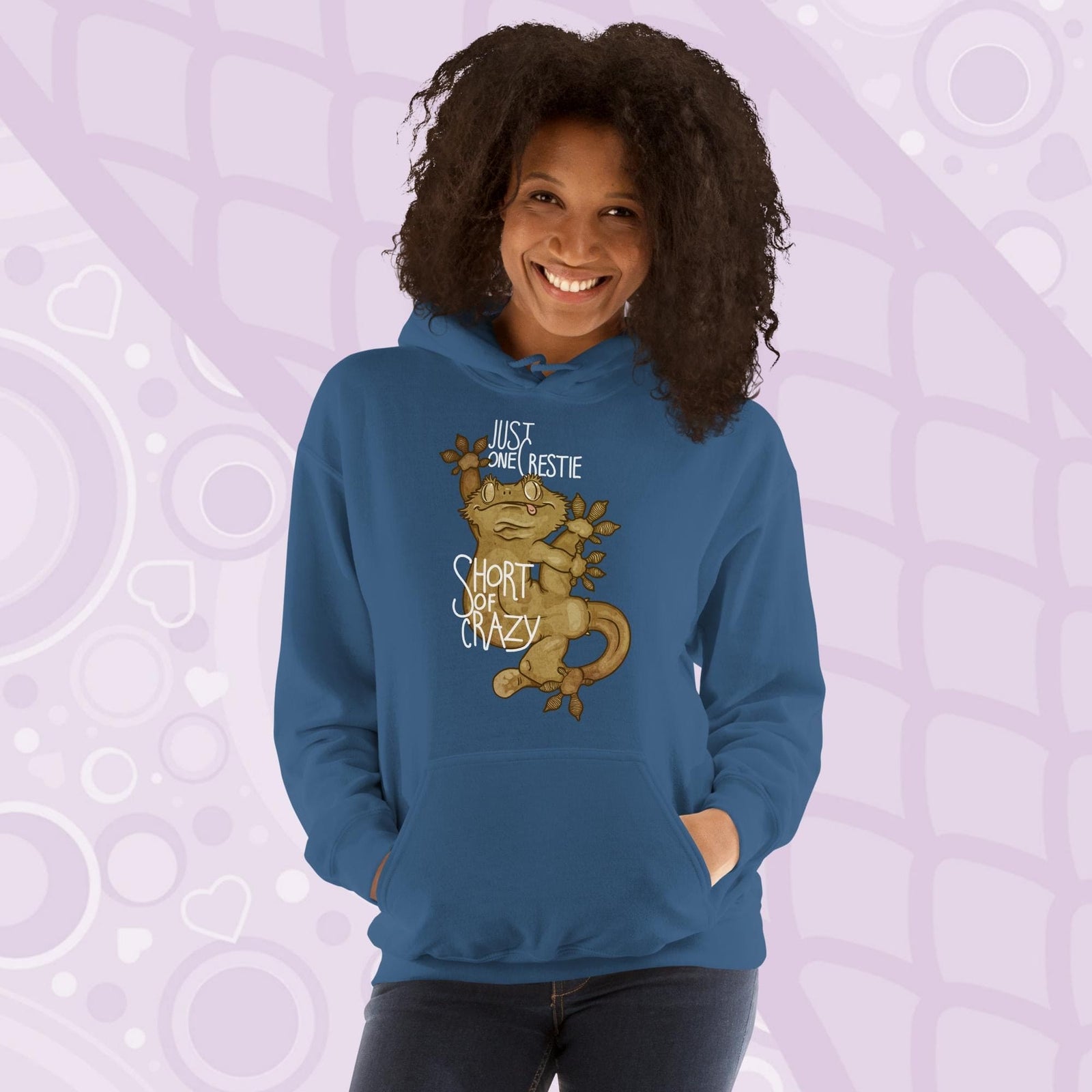 Woman wearing a blue colored pullover hoodie with a goofy, cartoonish graphic of a crested gecko with it's legs contorted in many directions. The text, "Just one crestie short of crazy." is printed on the front.