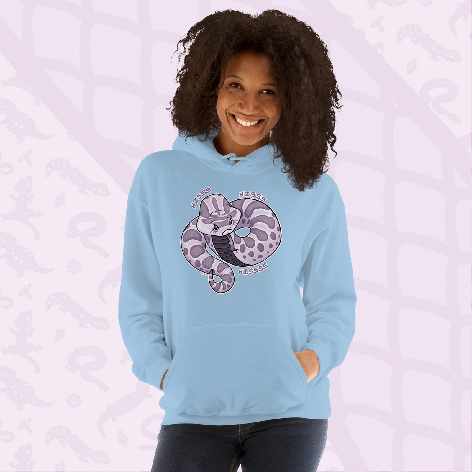 A woman wearing a soft light blue hoodie featuring a cartoon hognose snake design in shades of purple and gray. The snake is coiled and has the word "Hiss Hiss Hiss" printed around it. The hoodie has a front pocket and a hood.