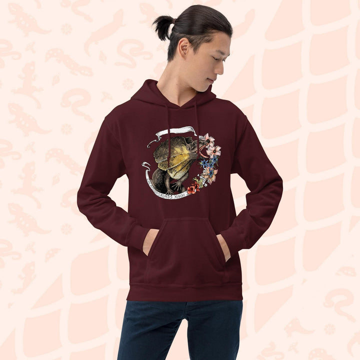 Man wearing a dark maroon red hooded sweatshirt with a graphic of a frilled lizard, it's mouth open and displaying it's pink and orange frills with a white banner framing it's shape. Pink flowers flank the right side of the design and the text "Chlamydosaurus Kingii" is along the bottom, which is the species name for frilled lizard. The hoodie has a front pocket.