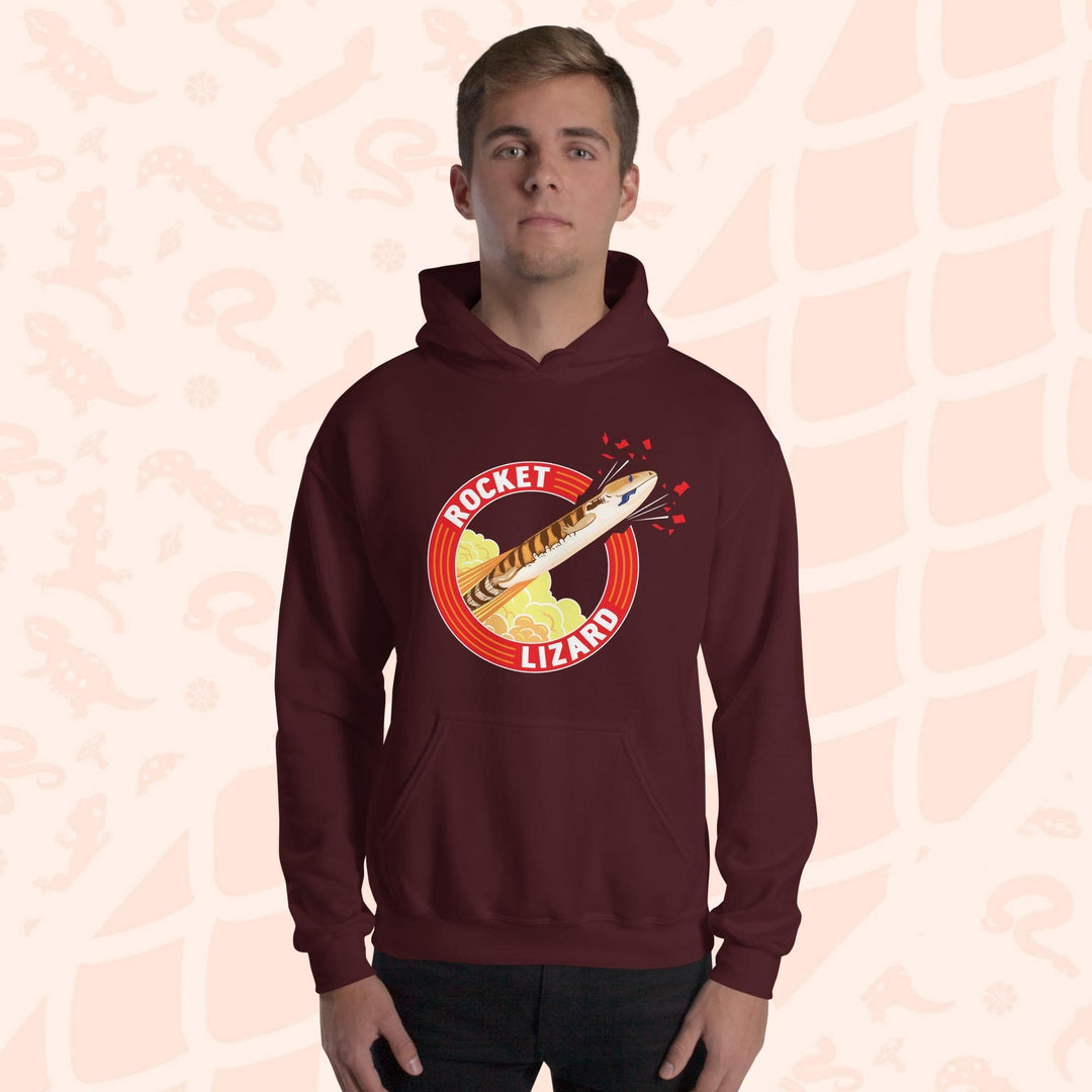 A man wearing a deep red maroon hooded sweatshirt with a red circle graphic with text saying "Rocket Lizard." In the center of the graphic is a cartoon blue tongue skink shooting upward and breaking through the red outer circle with the text. It's tongue is hanging out of it's mouth as it rockets out with a plume of yellow and orange clouds billowing behind it. The hoodie has a front pocket.