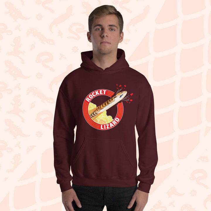 A man wearing a deep red maroon hooded sweatshirt with a red circle graphic with text saying "Rocket Lizard." In the center of the graphic is a cartoon blue tongue skink shooting upward and breaking through the red outer circle with the text. It's tongue is hanging out of it's mouth as it rockets out with a plume of yellow and orange clouds billowing behind it. The hoodie has a front pocket.