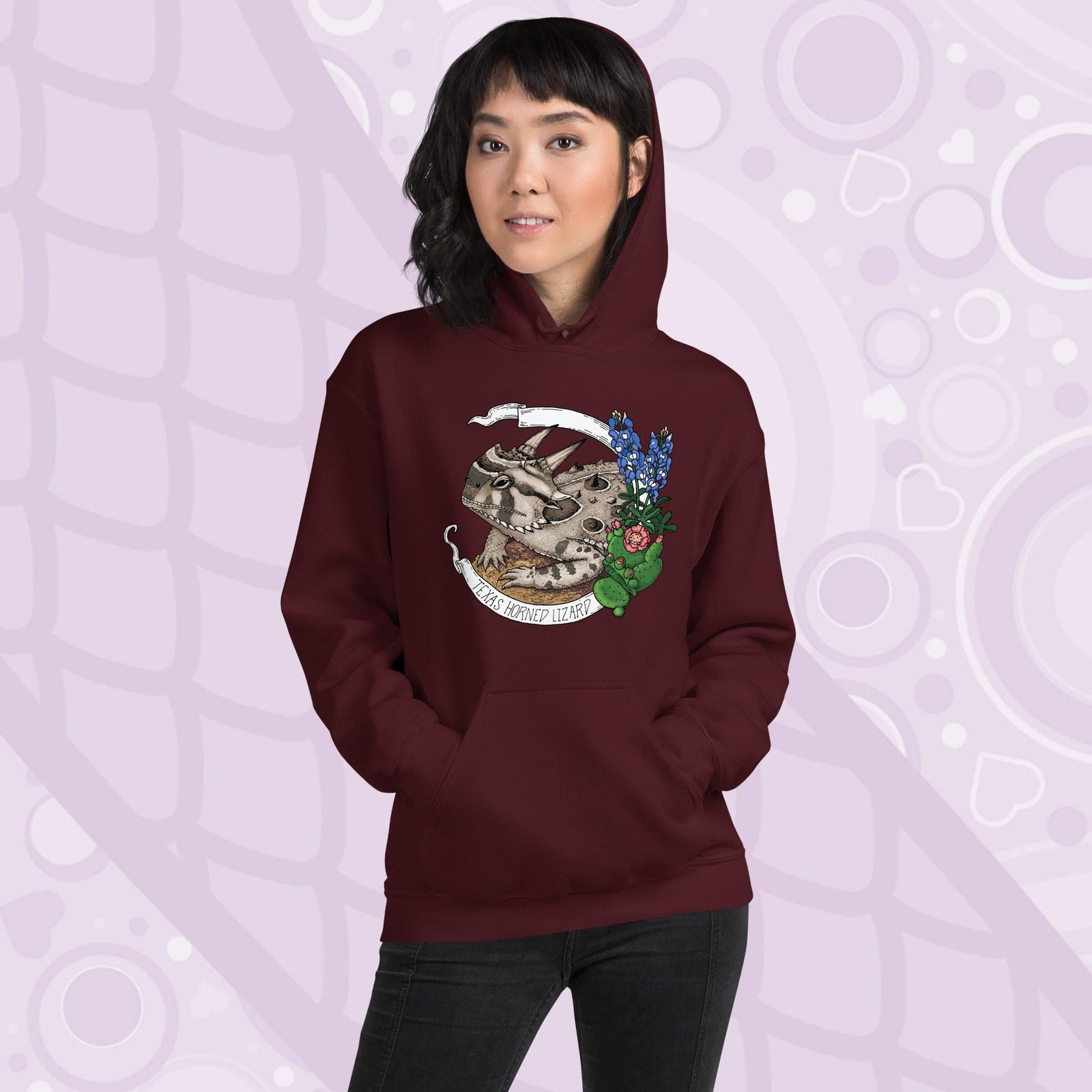A woman wearing a dark maroon red hoodie with a graphic of a Texas horned lizard surrounded by bluebonnets and a cactus. The text "Texas Horned Lizard" is printed in a white banner. The hoodie has a pocket in the front.