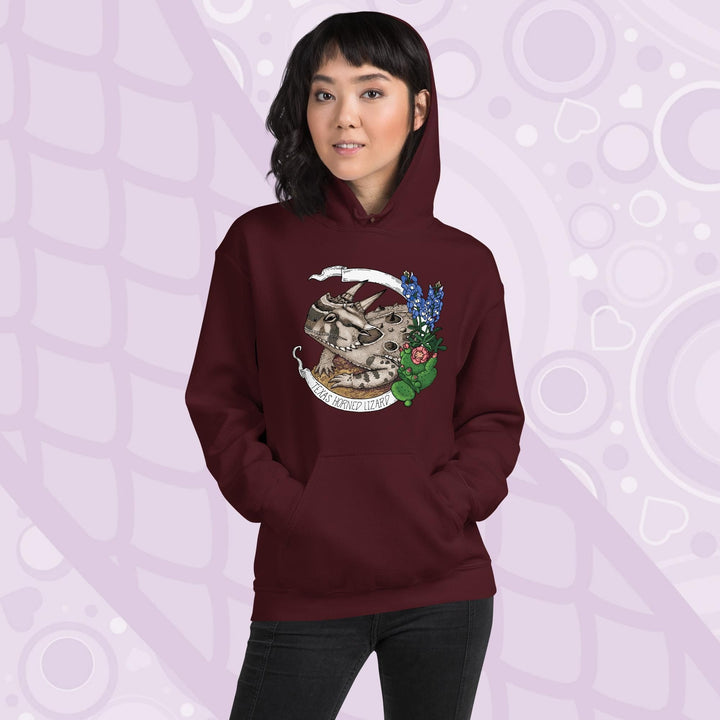 A woman wearing a dark maroon red hoodie with a graphic of a Texas horned lizard surrounded by bluebonnets and a cactus. The text "Texas Horned Lizard" is printed in a white banner. The hoodie has a pocket in the front.