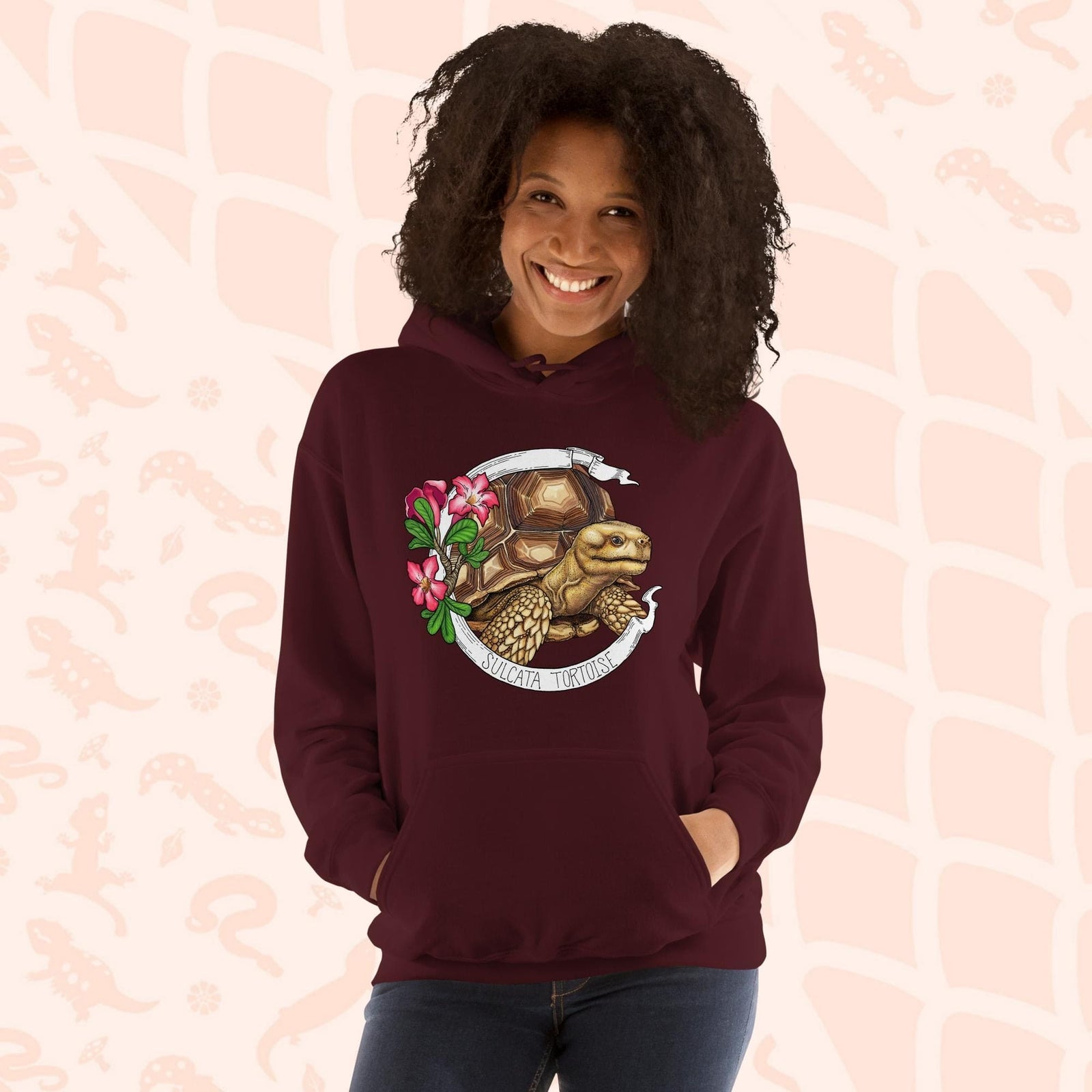 A woman wearing a dark maroon red hooded sweatshirt with a graphic of a Sulcata tortoise with large pink flowers on the left and a white banner framing above and below the artwork. Text "Sulcata Tortoise" is printed on the lower part of the banner. The hoodie has a front pouch.