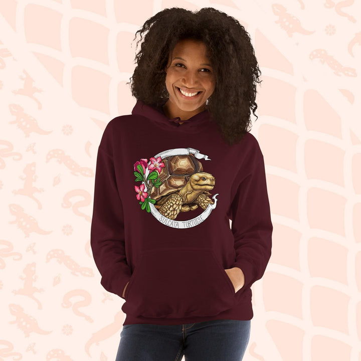 A woman wearing a dark maroon red hooded sweatshirt with a graphic of a Sulcata tortoise with large pink flowers on the left and a white banner framing above and below the artwork. Text "Sulcata Tortoise" is printed on the lower part of the banner. The hoodie has a front pouch.