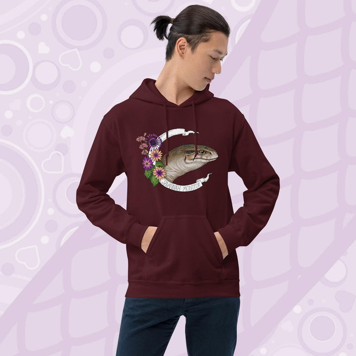 A  man wearing dark maroon red hooded sweatshirt with a graphic of an African Savannah monitor lizard with red flowers and the text "Savannah Monitor" on the bottom white banner that frames the reptile. The hoodie is a cotton fabric and has a front pouch.