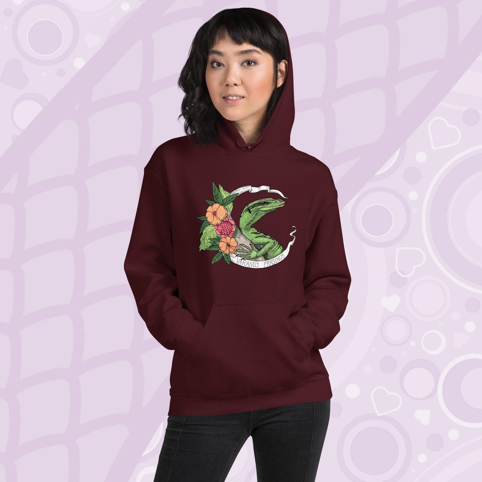 A woman wearing a deep maroon red hoodie with a graphic of a slender green tree monitor lizard with orange and warm orange red flowers and the text "Varanus Prasinus" on a white banner surrounding the reptile. The hoodie has a front pocket.