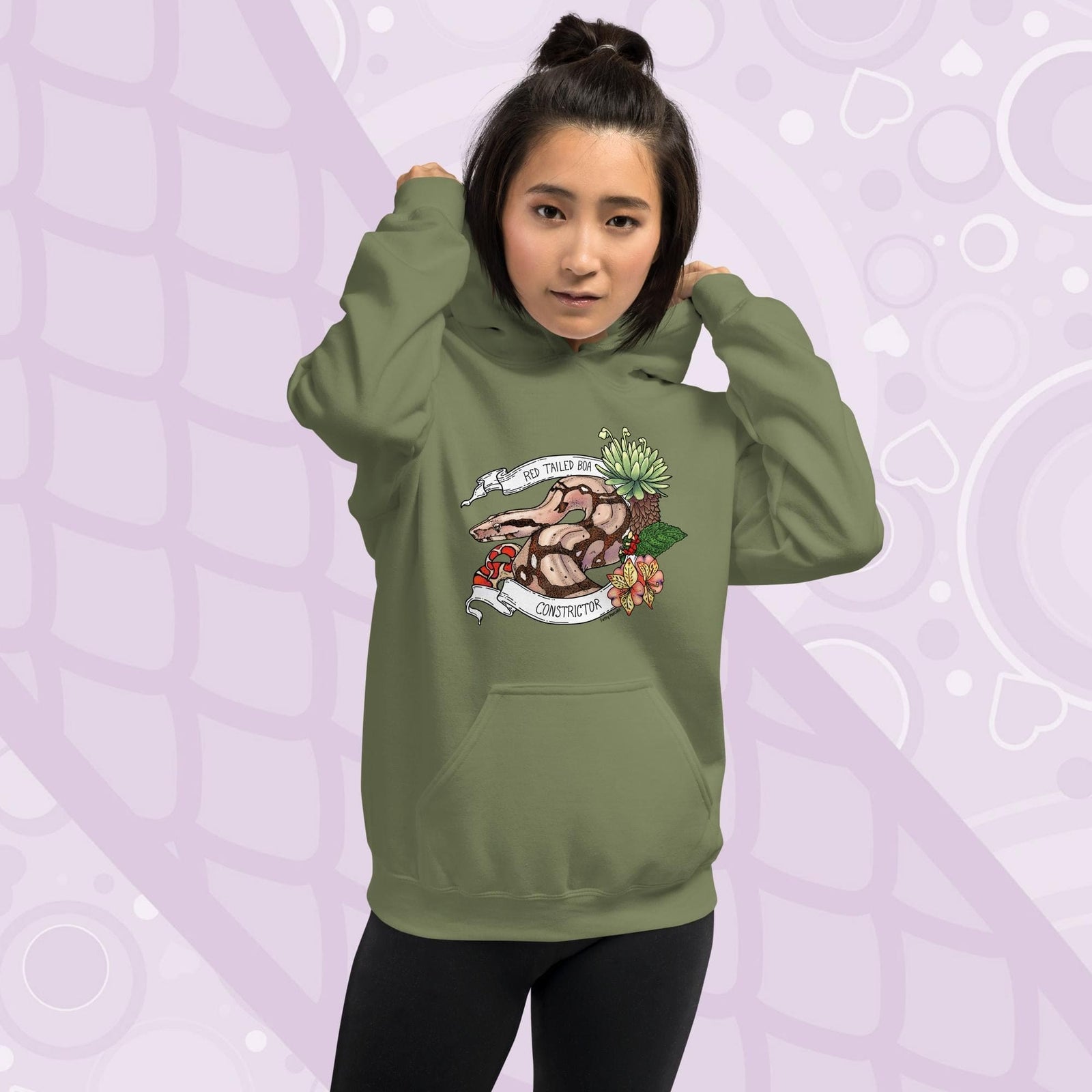A woman wearing a olive green pullover hoodie with a Red Tailed Boa Constrictor snake framed by a white banner and pink flowers with green foliage on the right side of the design. The hoodie is made of cotton and has a front pocket sewn in.