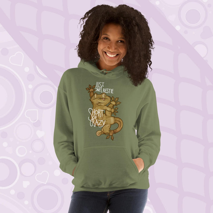 Woman wearing a olive green colored pullover hoodie with a goofy, cartoonish graphic of a crested gecko with it's legs contorted in many directions. The text, "Just one crestie short of crazy." is printed on the front.