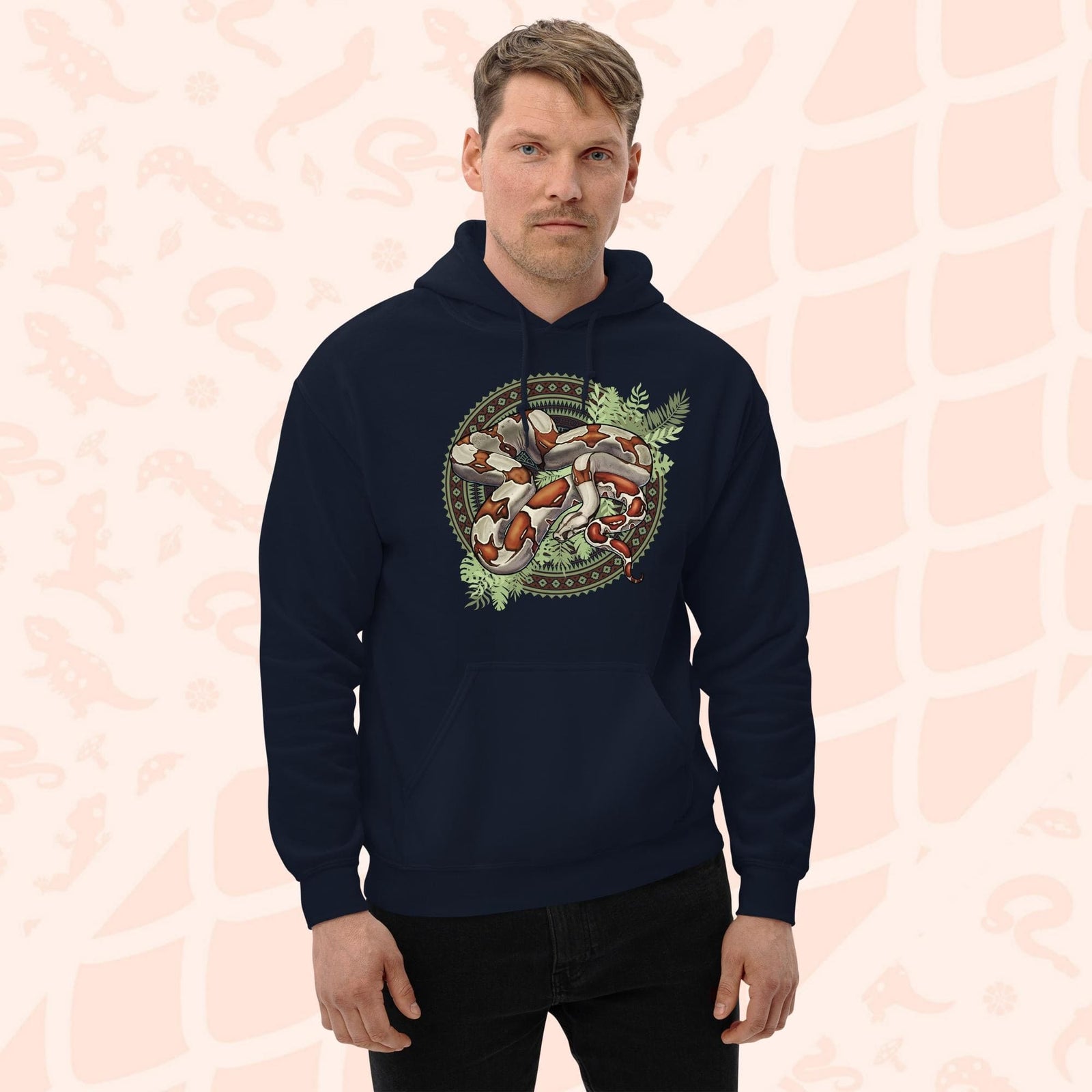 A man wearing a navy blue hooded sweatshirt with a graphic print of a red tailed boa constrictor snake with bold red's and brown markings. The snake is curled up in a circle made of patterned geometric shapes and with leafy green foliage beneath the snakes body. The hoodie has a front pocket.