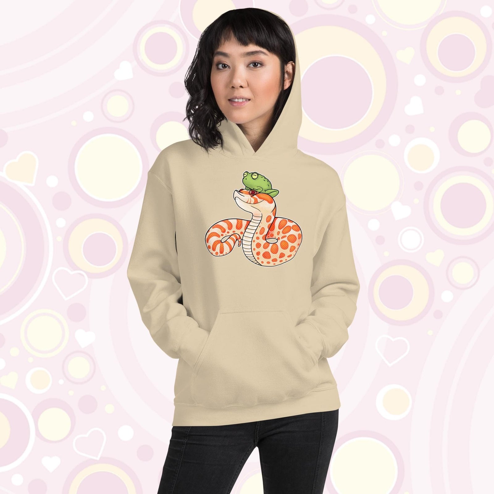 A woman wearing a soft cream pullover hoodie with a cartoon illustration of a albino hognose snake. It has bright orange and white spotted patterns and a silly frog sits on it's head. The hoodie is made of cotton and has a front pocket sewn in.