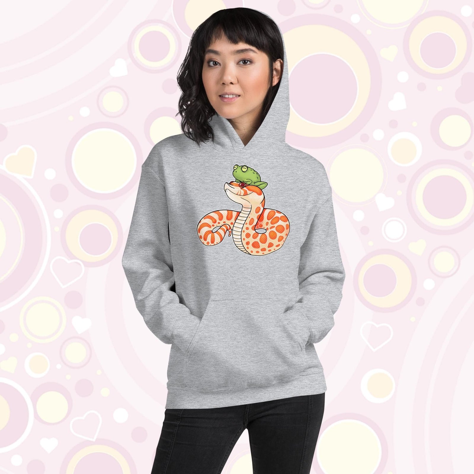A woman wearing a grey pullover hoodie with a cartoon illustration of a albino hognose snake. It has bright orange and white spotted patterns and a silly frog sits on it's head. The hoodie is made of cotton and has a front pocket sewn in.