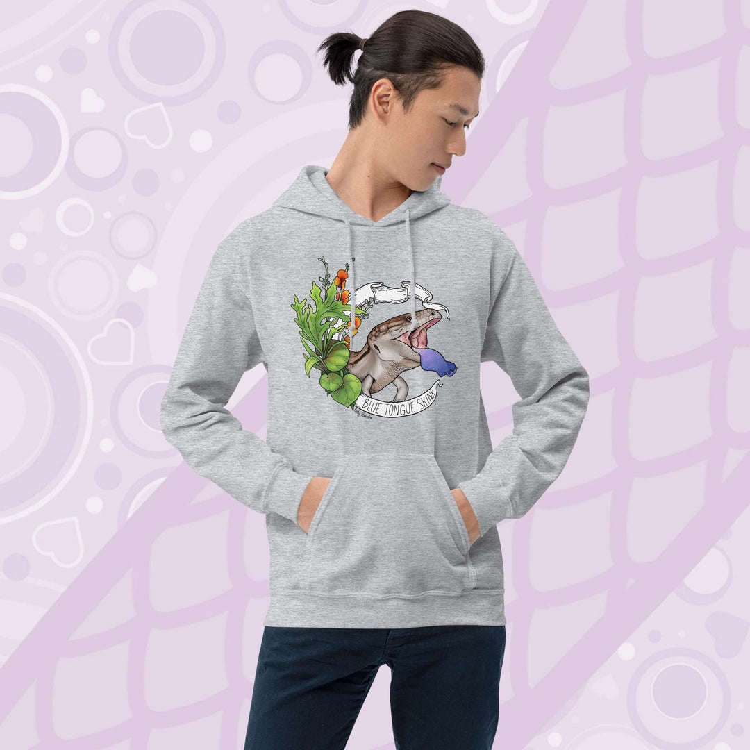 Man wearing a light grey hooded pullover sweatshirt with a graphic of a blue-tongued skink, it's mouth open and it's blue tongue flashing in a classic defensive posture. The lizard is surrounded by green leaves and flowers. The text "Blue Tongue Skink" is written on a white banner.