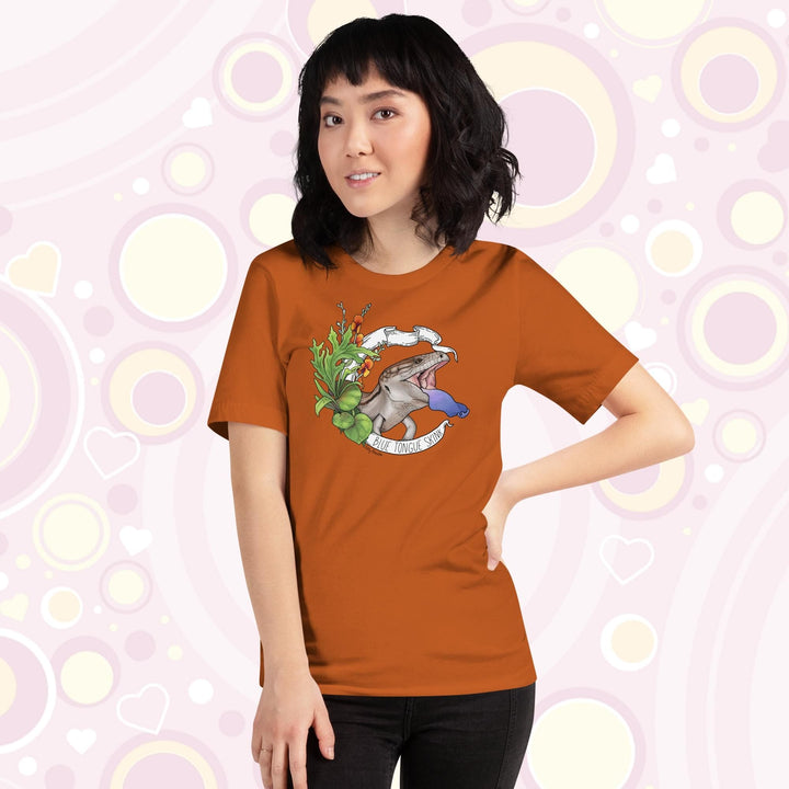 Lady wearing orangish colored tee with a hand drawn design of a blue tongue skink flashing it's blue tongue in warning. Framed with greenery and a white banner that reads, "Blue Tongue Skink".