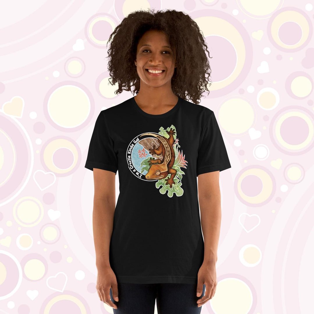 A woman wearing a black crew neck tee with a graphic of a crested gecko climbing down from a tree with the words "I'm a crestie kind of gal" on the left hand side.