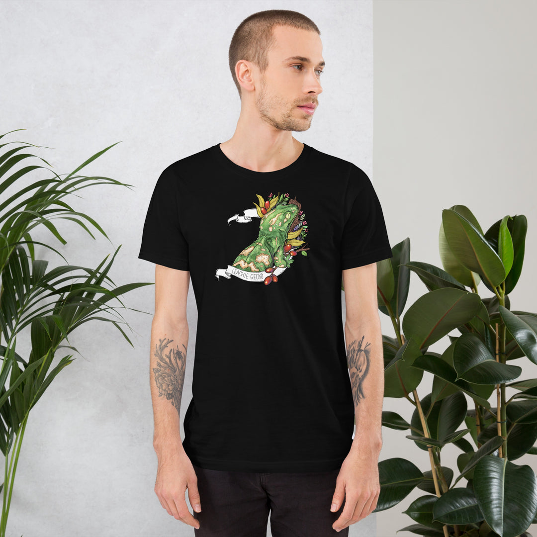 Man wearing a black t-shirt with a graphic of a leachie gecko on it, standing against a white wall with plants.