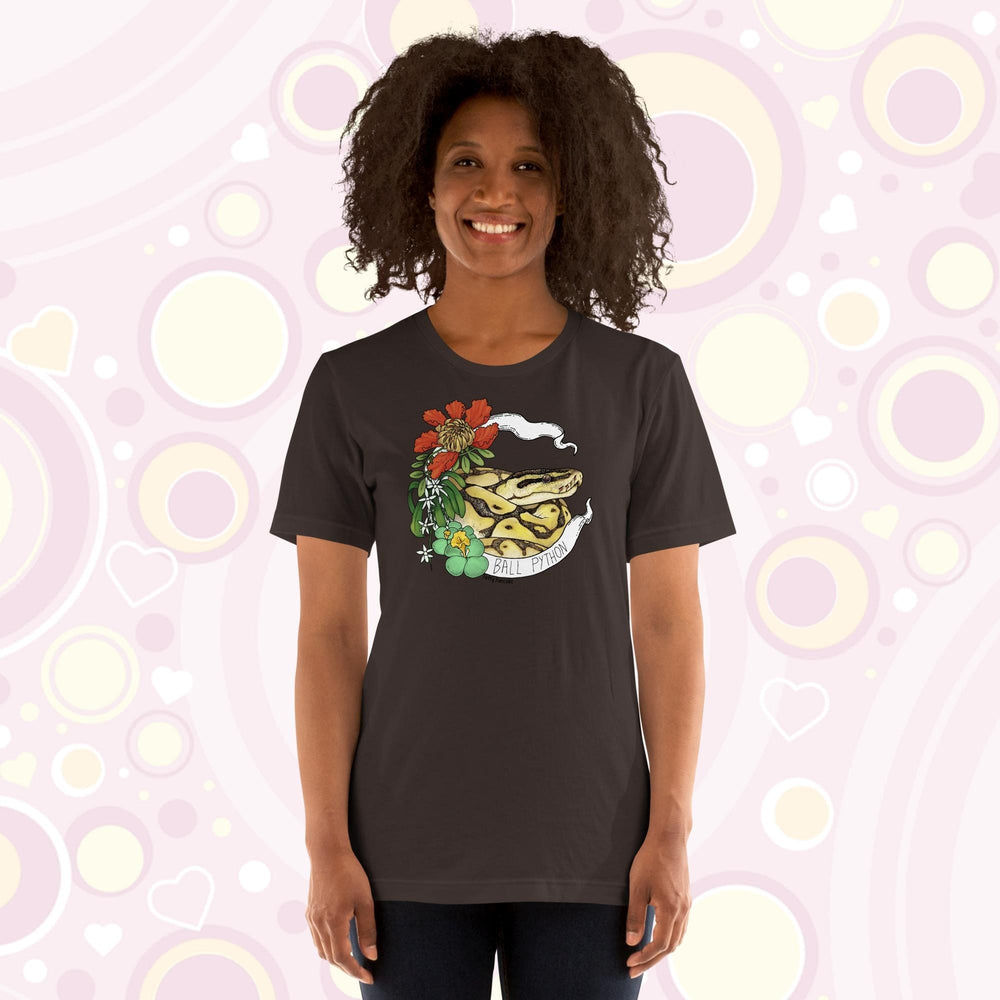 Image shows a woman wearing a brown crew neck tee with a ball python coiled up in a white banner with the snake species on it. Red, white, and green flowers and plants adorn the left side of the graphic.