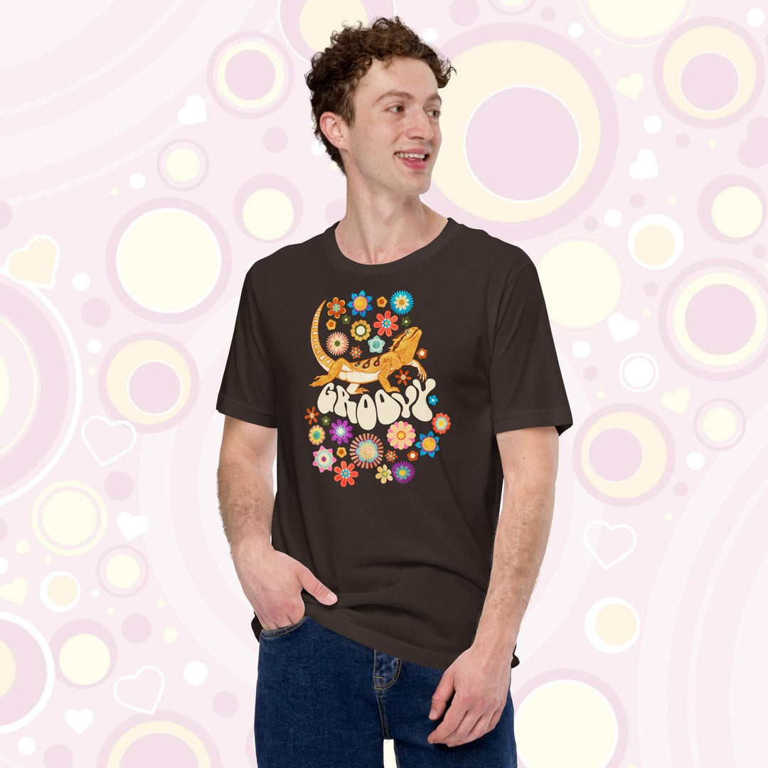 A young man wearing a brown crew neck tee showing a groovy bearded dragon design with a 70's retro style with many multi colored flowers surrounding a strutting bearded dragon lizard with the text "groovy" under it's body.